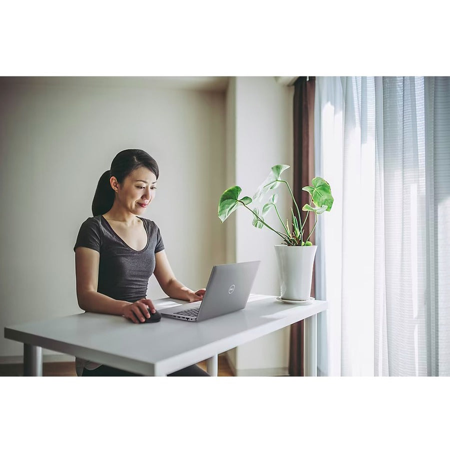 Woman using a Dell laptop with the Dell MS300 Wireless Mouse, showcasing a modern, productive workspace with a plant and natural light.